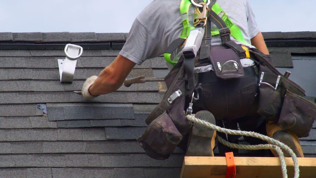 A roofing contractor in safety gear repairing asphalt shingles on a residential roof using professional tools.