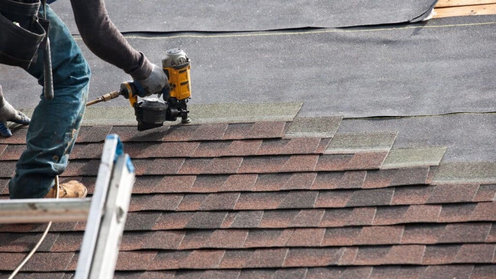 Close-up of roofing installation with a professional using a nail gun on asphalt shingles.