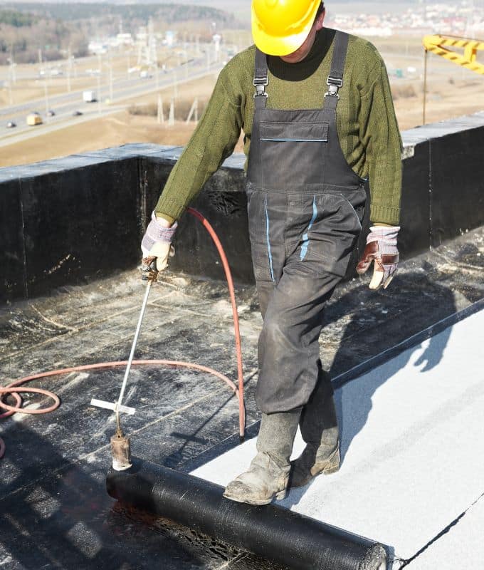 Roofer applying a torch-down roofing membrane on a flat commercial roof for waterproofing.