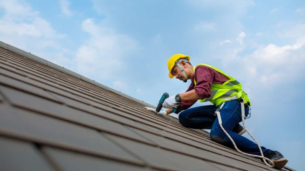 Contractor fixing a metal roof while safely secured with a harness under blue skies.