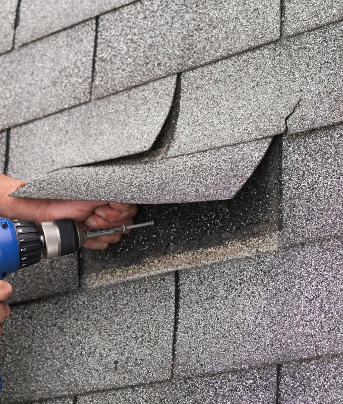 Close-up of a contractor using a drill to secure a nail under asphalt shingles during roof repair.