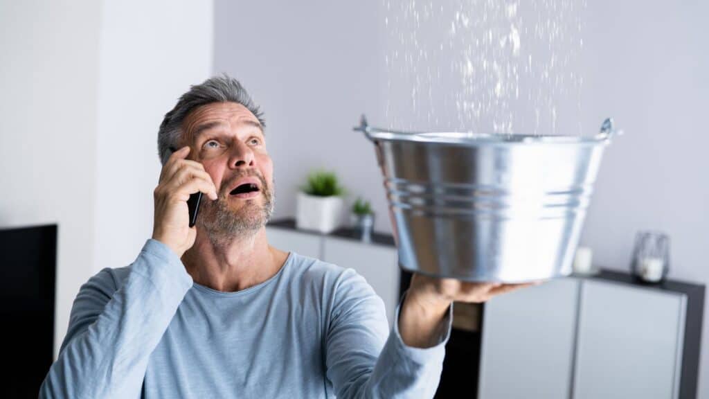 Man catching water in a bucket from a leaking ceiling, calling for Roof Repairs.